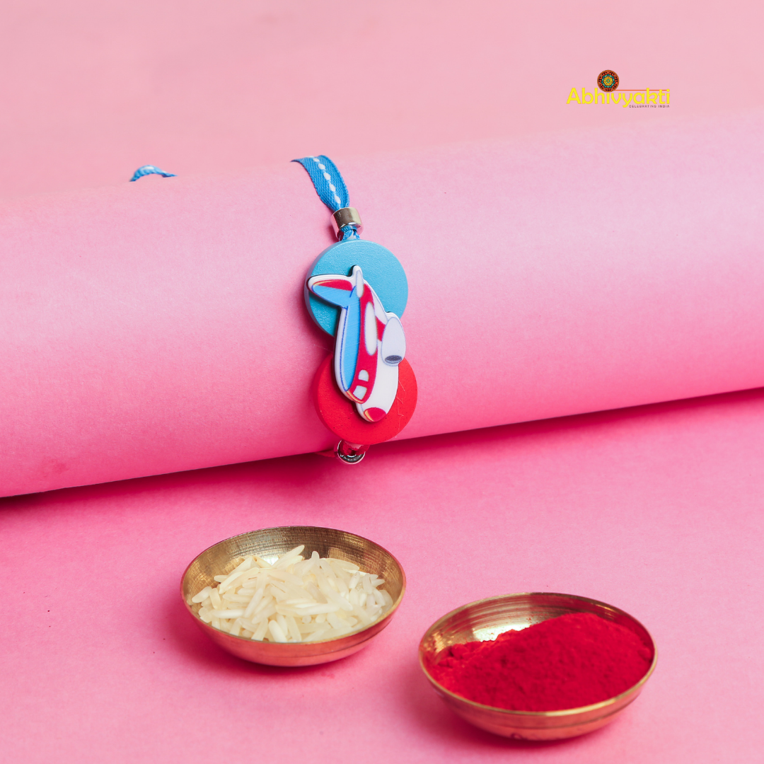 A colorful kids rakhi with a plane design on a pink background, placed near two brass bowls one with rice and the one with roli.