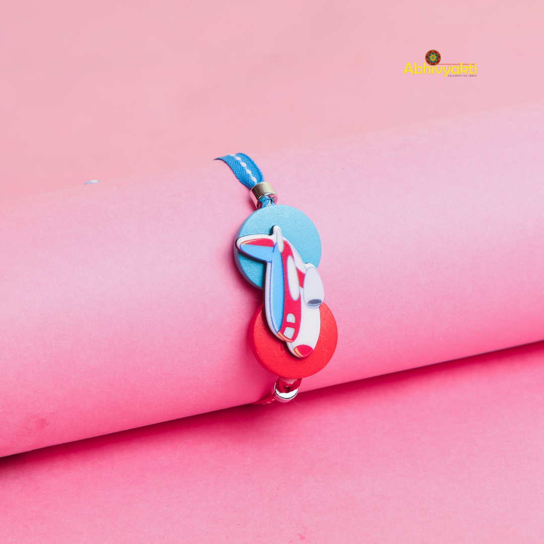 A blue and red Kids rakhi with a cartoon airplane charm and round beads is displayed on a rolled pink paper background. The Abhvyakti logo is in the top right corner.