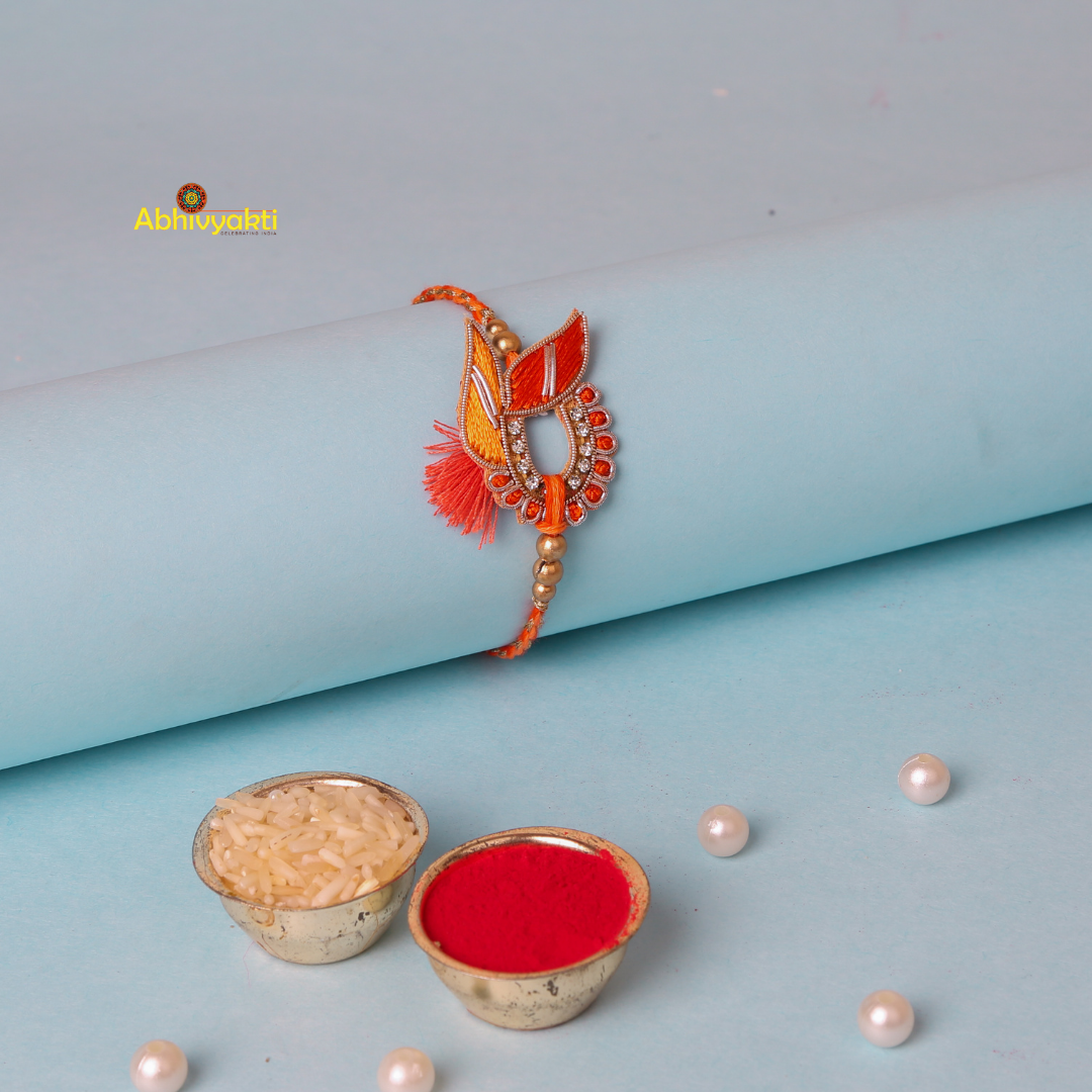 An artistic, orange and gold zari rakhi with beads and stone gracefully rests on a light blue cylindrical surface. The intricate design features leaf-like motifs, bead accents, and a central circular section adorned with small diamonds. A tassel detail adds a touch of elegance.