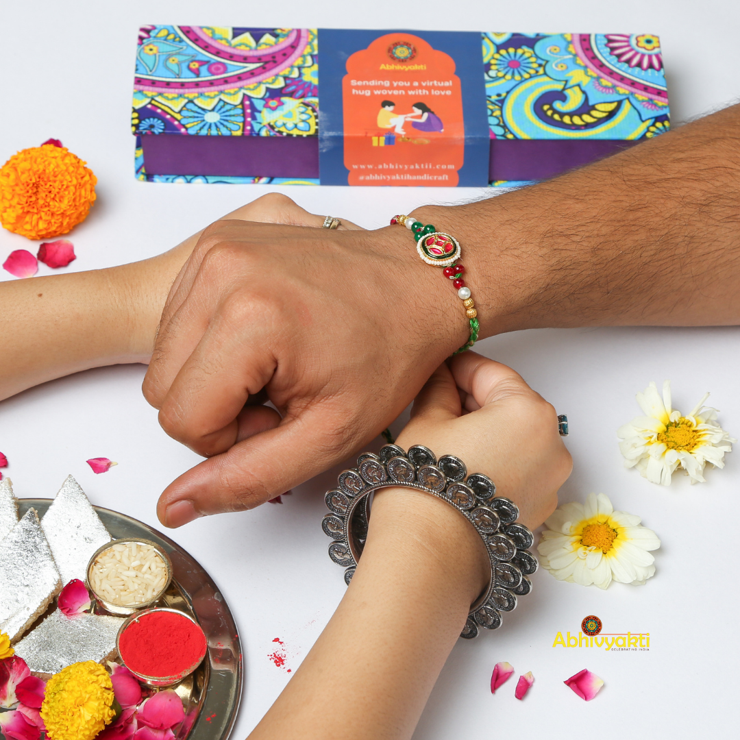 A beaded red and gold-plated rakhi against a light blue background. The rakhi features a central circular emblem with red and green intricate designs, flanked by beads of various colors including white, green, and gold, with a braided red and green thread.