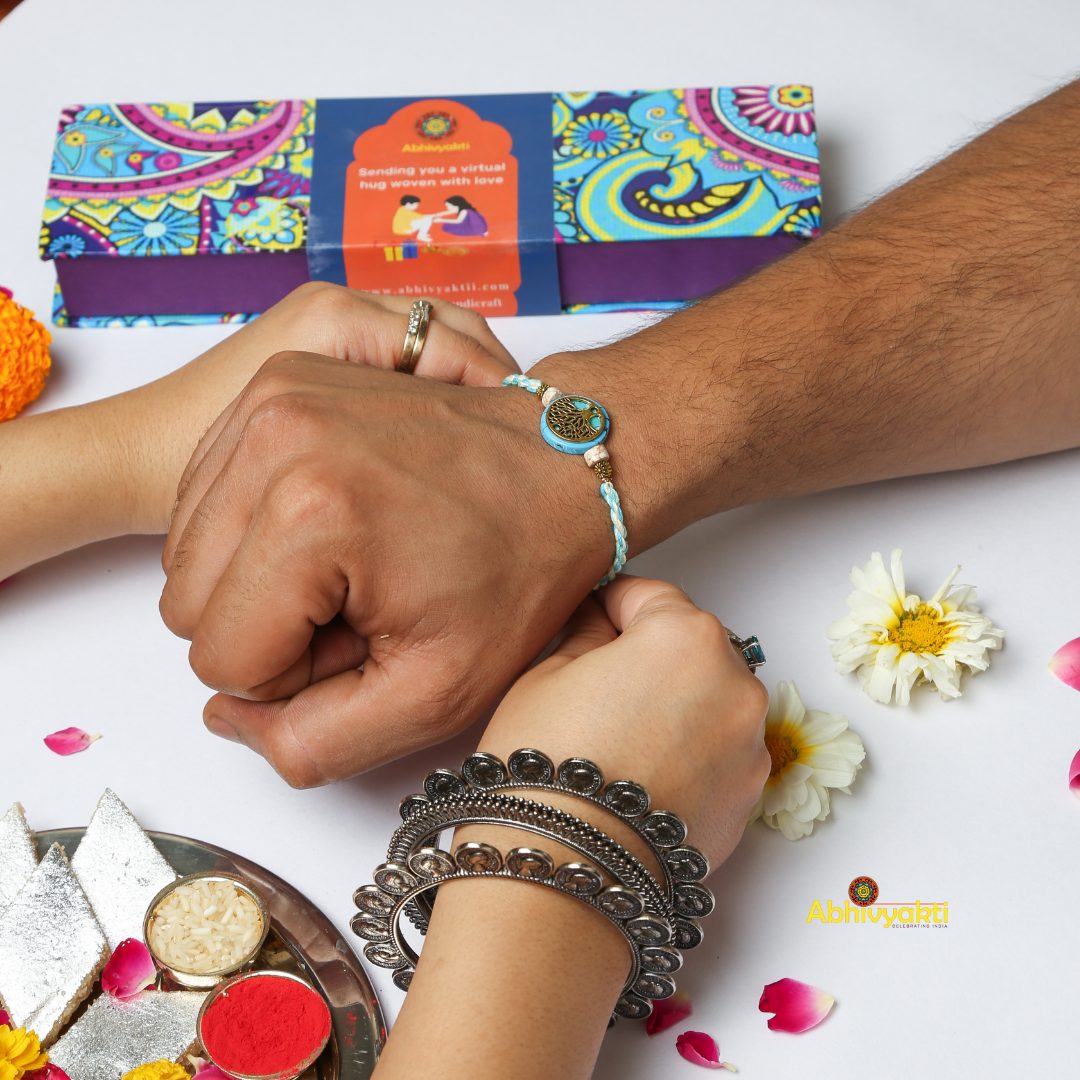 A braided bracelet with blue and white threads, featuring a circular charm with an intricate tree of life design in gold. The charm is flanked by two smaller pink beads and metallic accents, set against a pastel blue background—perfect as a gold plated rakhi for special occasions.