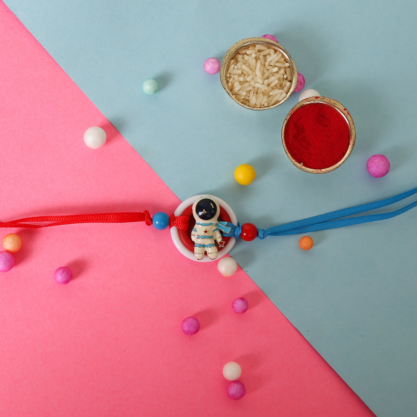 A colorful bracelet with a small astronaut charm lying on a pink surface, resembling a festive rakhi for kids. The bracelet features red and blue strings with round beads, and the astronaut is white with blue and red details, encircled by a red and white ring.