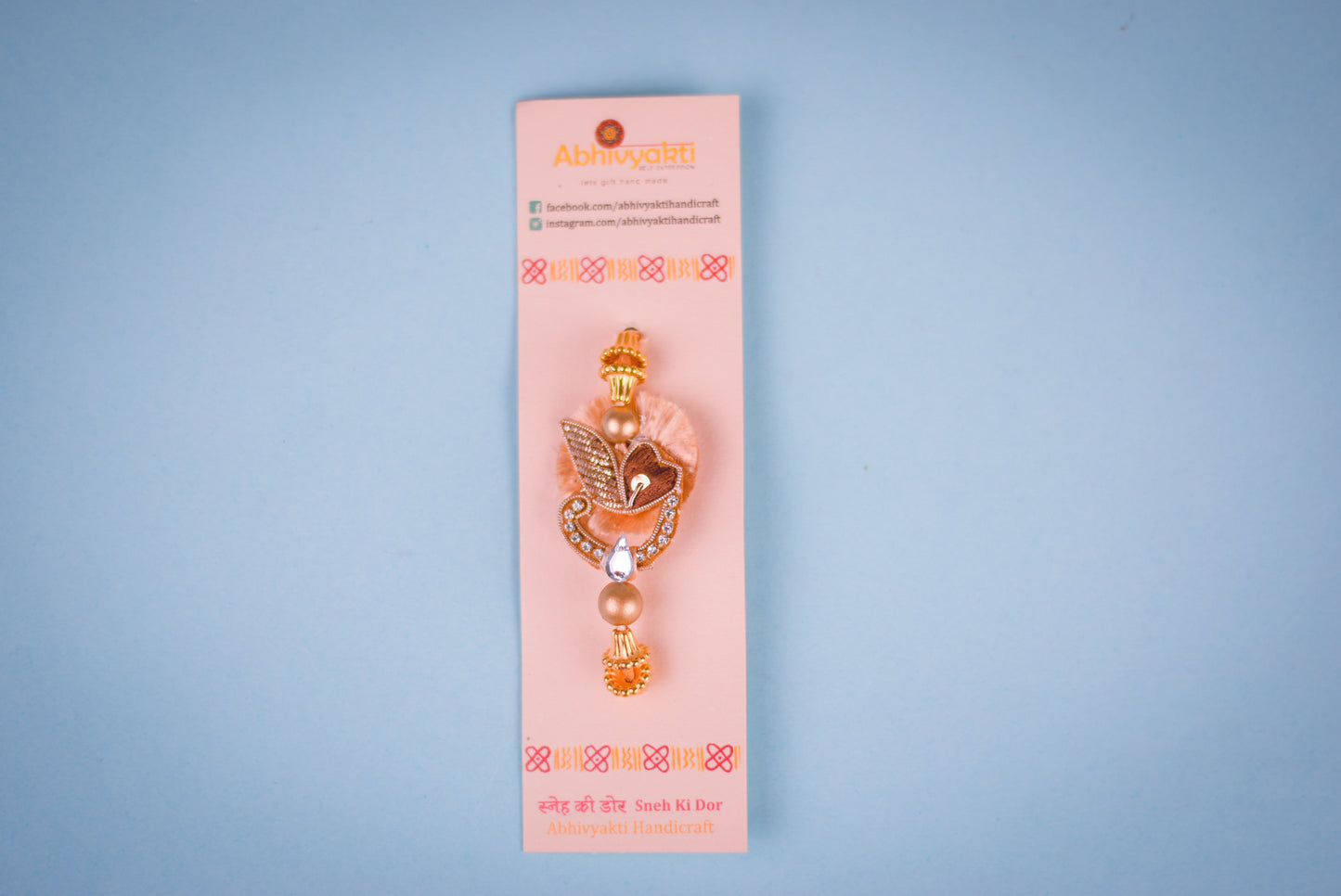 Fancy rakhi featuring a gold and brown leaf motif with sparkling stones, beige pom-pom, hanging bead, placed on a blue roll with rice, roli, and a pearl bracelet nearby