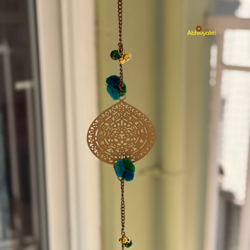 Filigree metal door hanging in a circular shape featuring detailed ornamental design, adorned with blue pom-poms and tiny golden bells, suspended from a chain indoors near a window