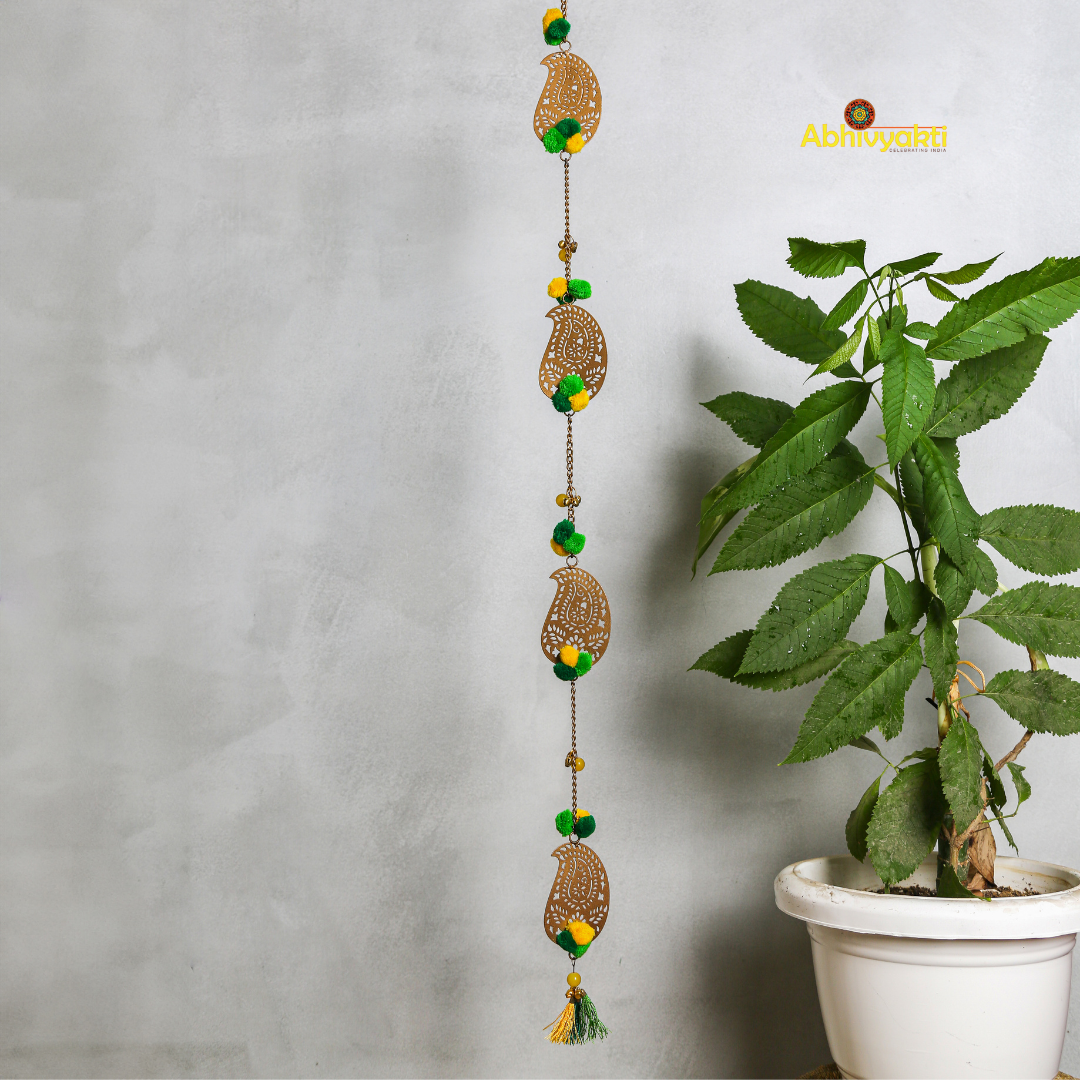 Filigree paisley-shaped metal door hanging adorned with yellow and green pom-poms and tassels, hanging on a white wall near a wooden door