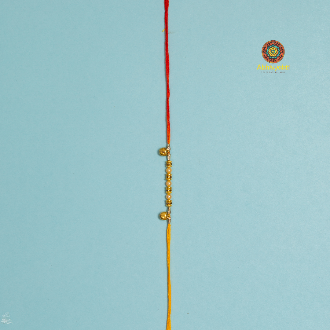 Golden bead and pearl rakhi on a yellow thread, wrapped around a blue roll, with bowls of rice and red kumkum powder, scattered pearls, thread rakhi with beads and ghungru.