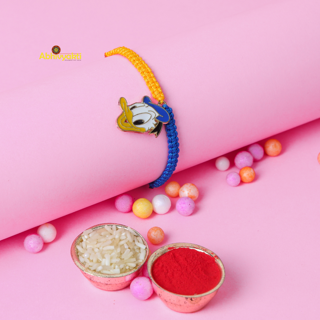 Kids rakhi featuring a Donald Duck cartoon charm on a dual colored braided thread (orange and blue) with colorful fabric balls scattered on a Pink background