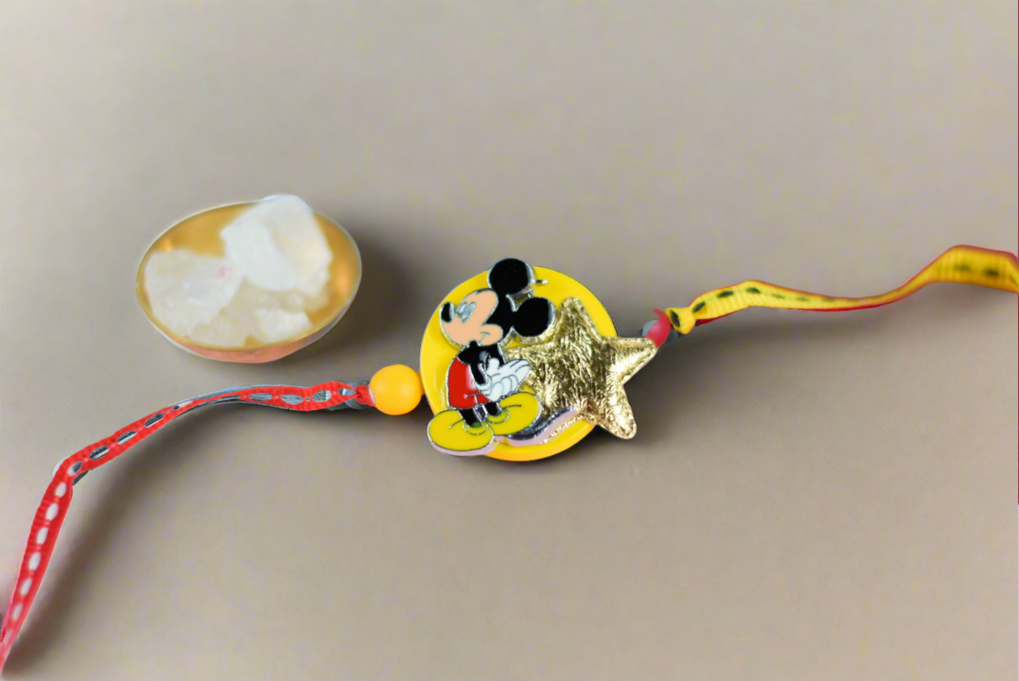 Kids rakhi featuring a cute Mickey Mouse charm and a golden star on a yellow base, tied with red and yellow thread and beads. Bowls of sugar, red roli, and rice are in the background on beige