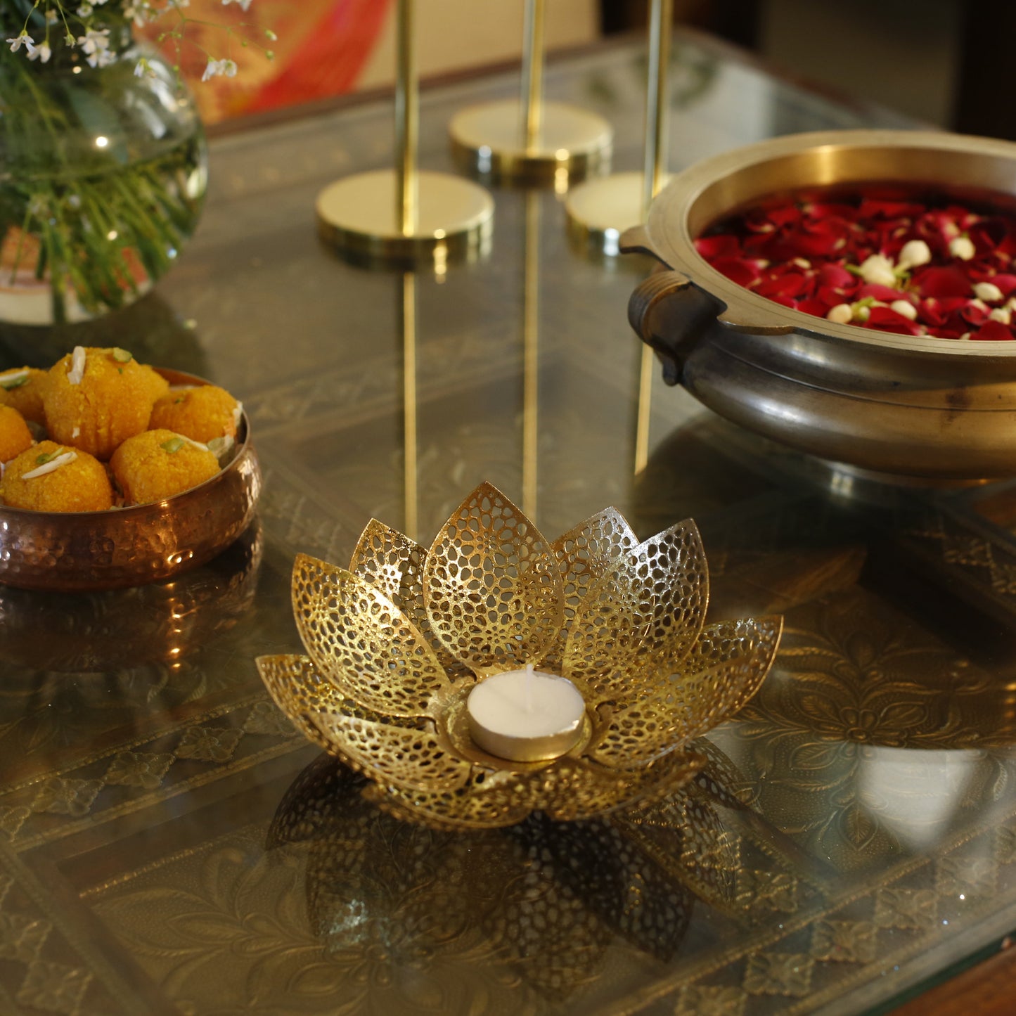 Decorative items including a gold lotus-shaped candle holder, small bowl with yellow items, and a pot with red flowers on a reflective surface.