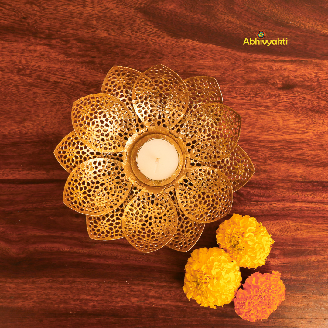 Gold-colored metal lotus flower-shaped t-light holder ,Padam on a wooden surface, with a white t-light candle inside, accompanied by artificial yellow and orange flowers to the side.