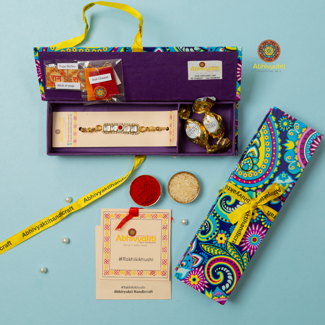 Traditional Rakhi with a rectangular gold centerpiece featuring three stones and a red stone in the center, decorated with gold beads and cream threads, placed on a light blue background with chocolates, roli and rice.