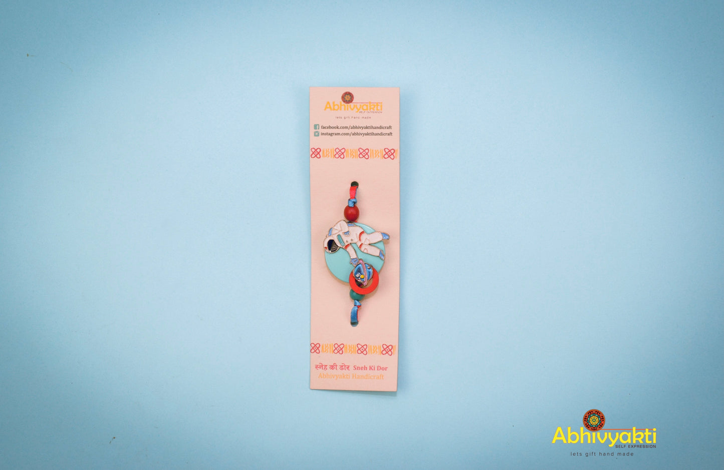 Kids rakhi with an astronaut charm reaching for a blue and red planet, set on a teal round base, tied with blue and red ribbon.