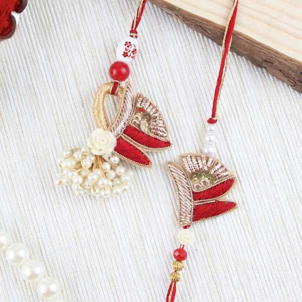 Decorative rakhi lumba set with red and gold fan shaped motifs, pearl clusters, and floral accents, displayed on a white hexagonal tile with rice, roli, sweets, and rose petals on a golden fabric background