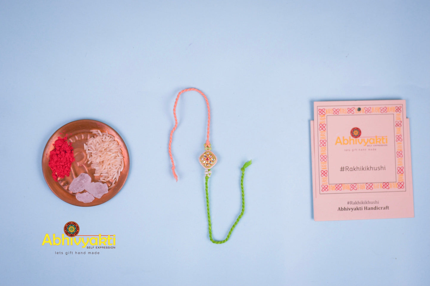 Elegant rakhi with a gold and green centerpiece, decorated with small pearls and colored stones, tied with green and peach threads, placed on a blue background with rice and kumkum