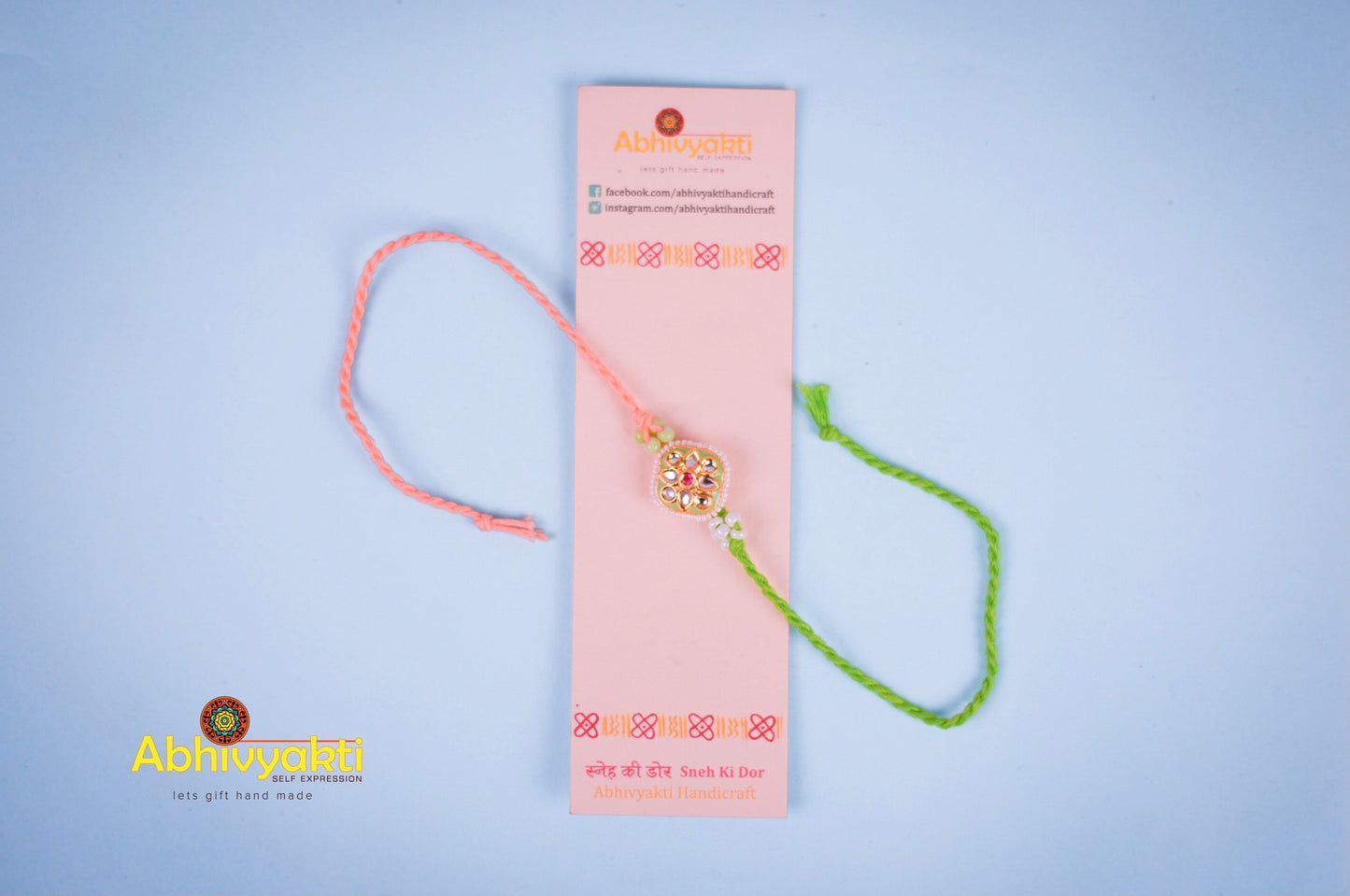 Elegant rakhi with a gold and green centerpiece, decorated with small pearls and colored stones, tied with green and peach threads, placed on a blue background