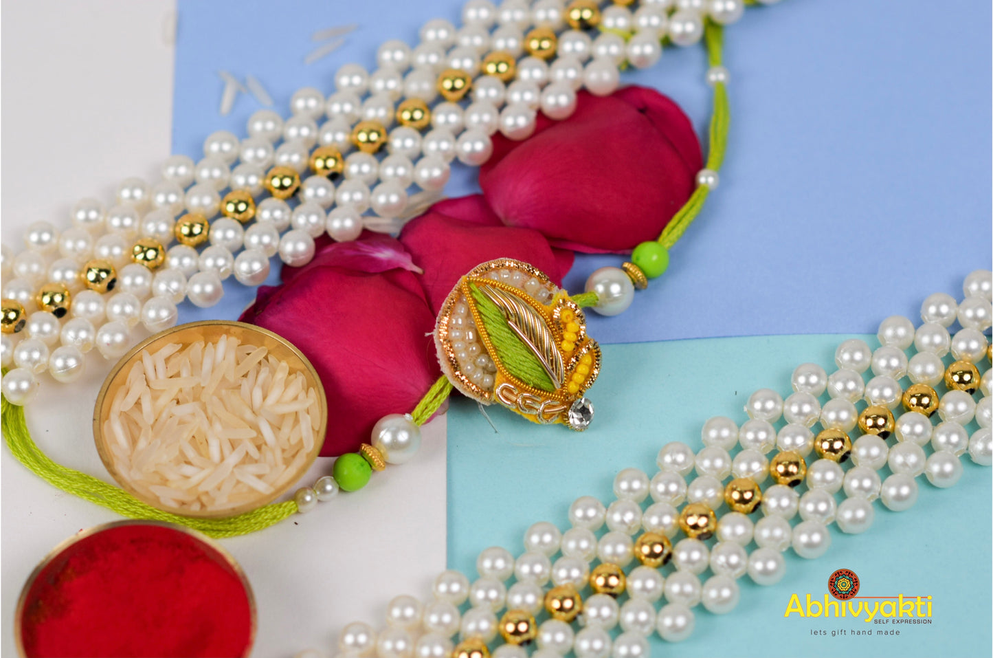 Green thread rakhi with a gold and green leaf centerpiece, pearl and green beads, placed on a blue roll with rice, roli, a Ganesh idol, and a pearl bracelet