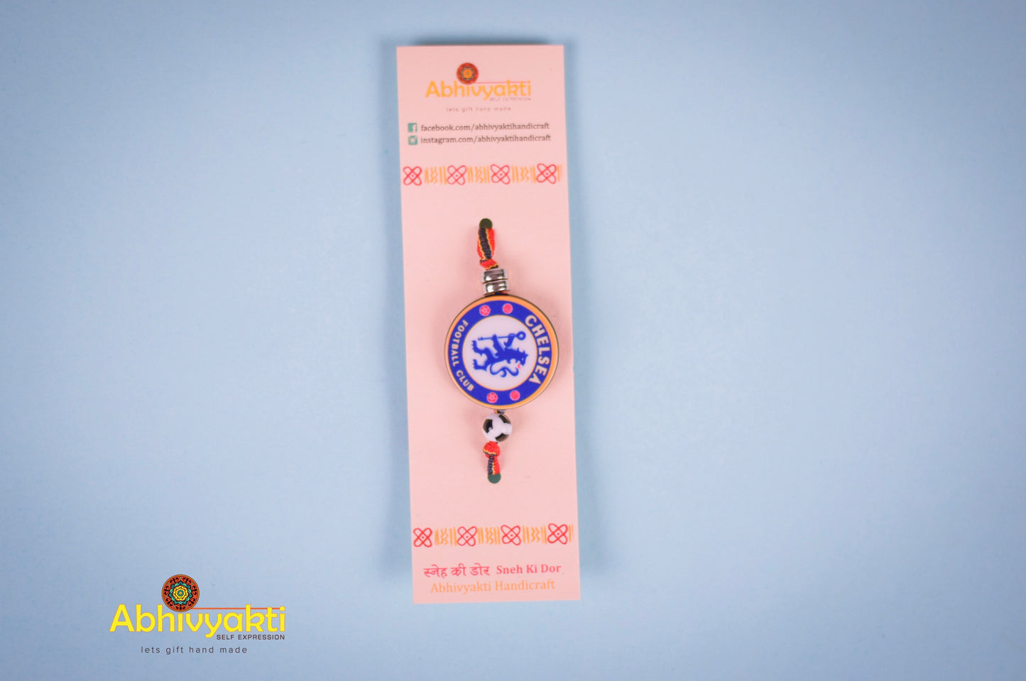 Kids rakhi featuring a Chelsea Football Club badge at the center, tied with a red, yellow, and blue striped thread and a football bead. Bowls of sugar, red roli, and rice are in the background on a blue surface