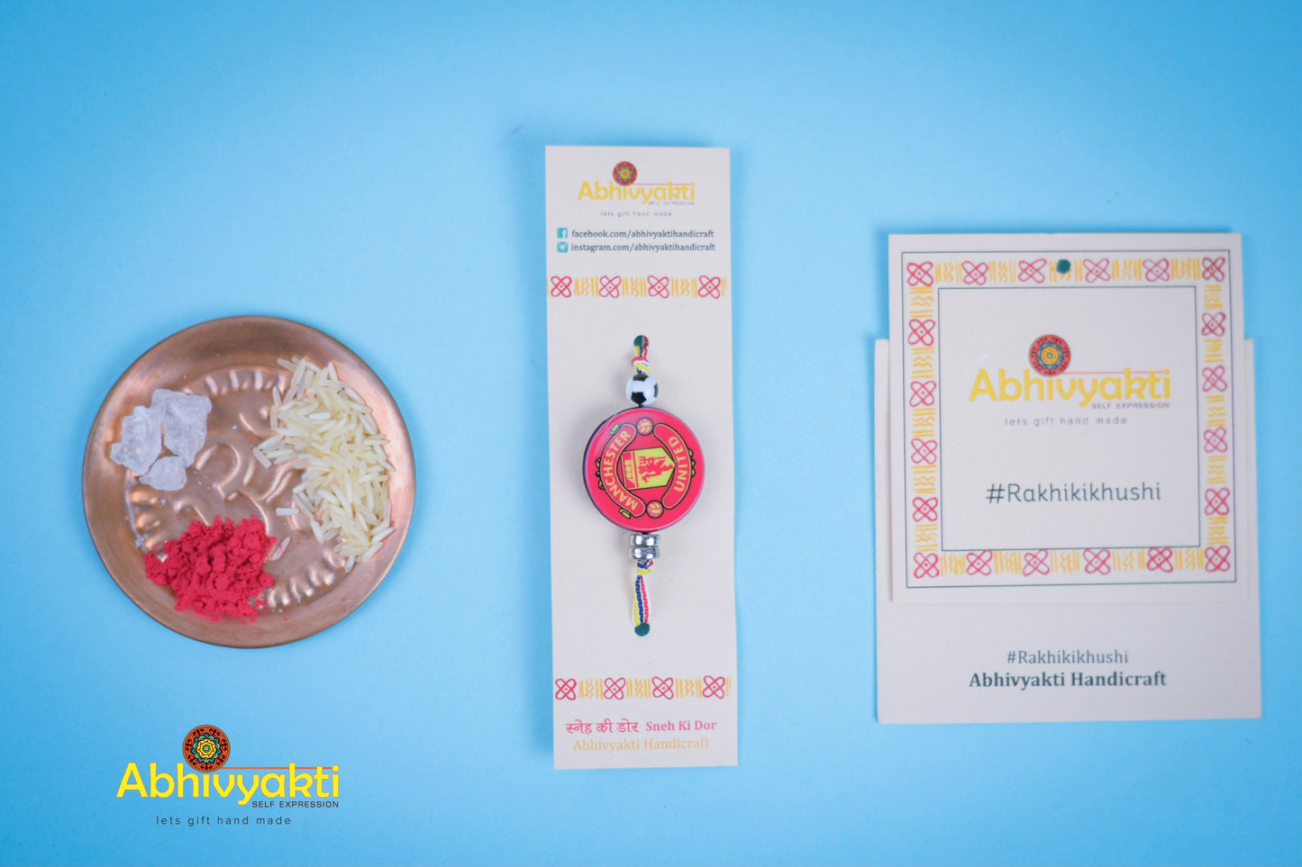 Kids rakhi featuring a Manchester United badge at the center, tied with a red, yellow, and white striped thread and a small football bead. Bowls of sugar, red roli, and rice are in the background on a blue surface