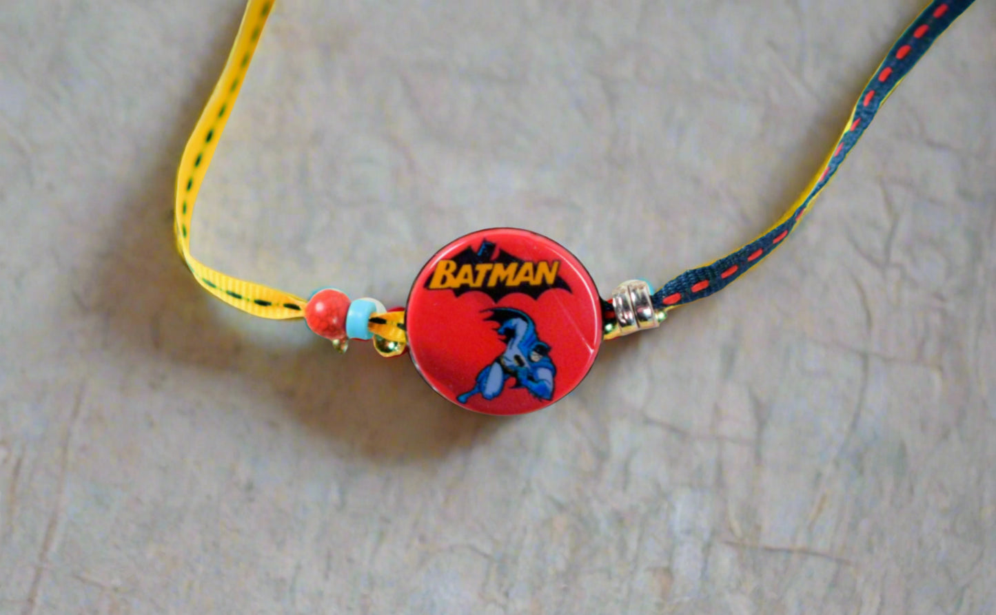 Kids rakhi with a red round Batman badge at the center, featuring Batman’s logo and character, tied with yellow and blue stitched thread and colorful beads, on a plain beige background