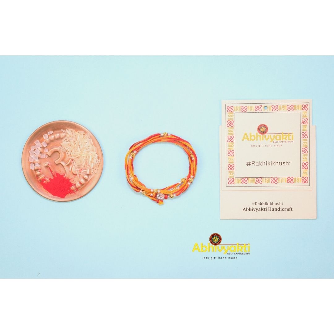 Simple stone rakhis with red and yellow threads and small gold beads, one with a clear stone in the center, all wrapped around a blue roll. Bowls of rice and roli, scattered pearls