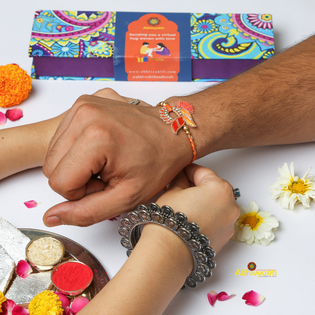 An artistic, orange and gold zari rakhi with beads and stone gracefully rests on a light blue cylindrical surface. The intricate design features leaf-like motifs, bead accents, and a central circular section adorned with small diamonds. A tassel detail adds a touch of elegance.