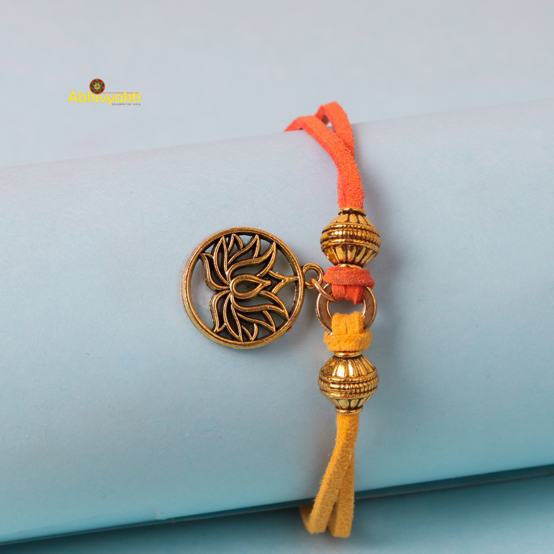 A decorative gold plated rakhi with a round lotus design in metallic gold, adorned with two additional gold beads. The rakhi is tied with vibrant orange and yellow threads, placed against a light blue background.