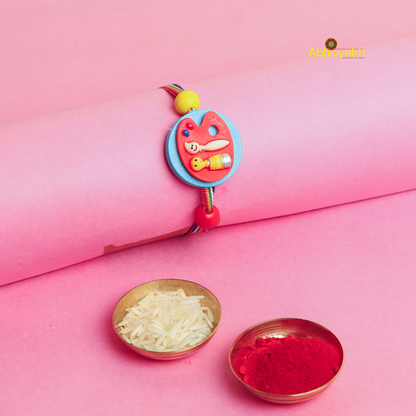 A decorative rakhi with a playful design is placed on a pink background near two small bowls, one filled with rice and the other with Roli. 