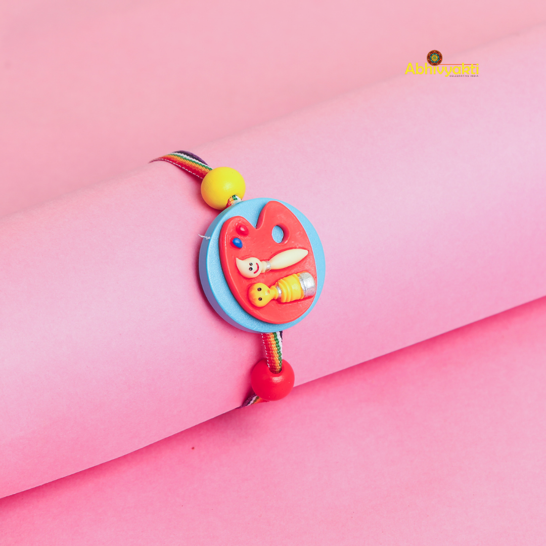 A colorful rakhi with a painter’s palette charm, brush, and paint tube decor on a multicolored thread, displayed on a pink background.