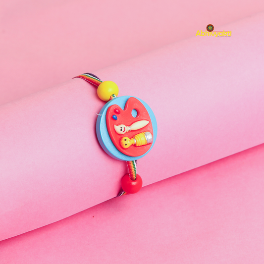 A colorful rakhi with a painter’s palette charm, brush, and paint tube decor on a multicolored thread, displayed on a pink background.