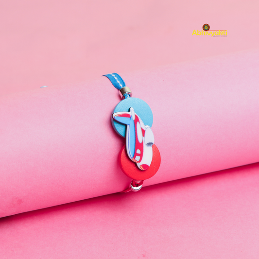 A blue and red Kids rakhi with a cartoon airplane charm and round beads is displayed on a rolled pink paper background. The Abhvyakti logo is in the top right corner.