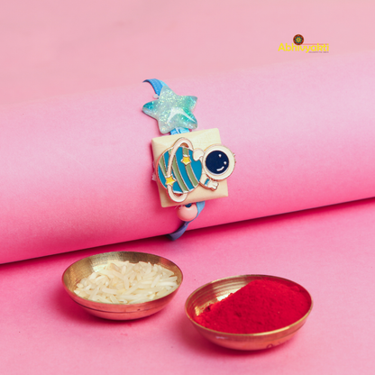 Cartoon astronaut and planet Rakhi bracelet for kids, featuring a blue strap, a glittery star, and a cute astronaut with a planet charm, with two bowls rice and roli, displayed on a pink background.