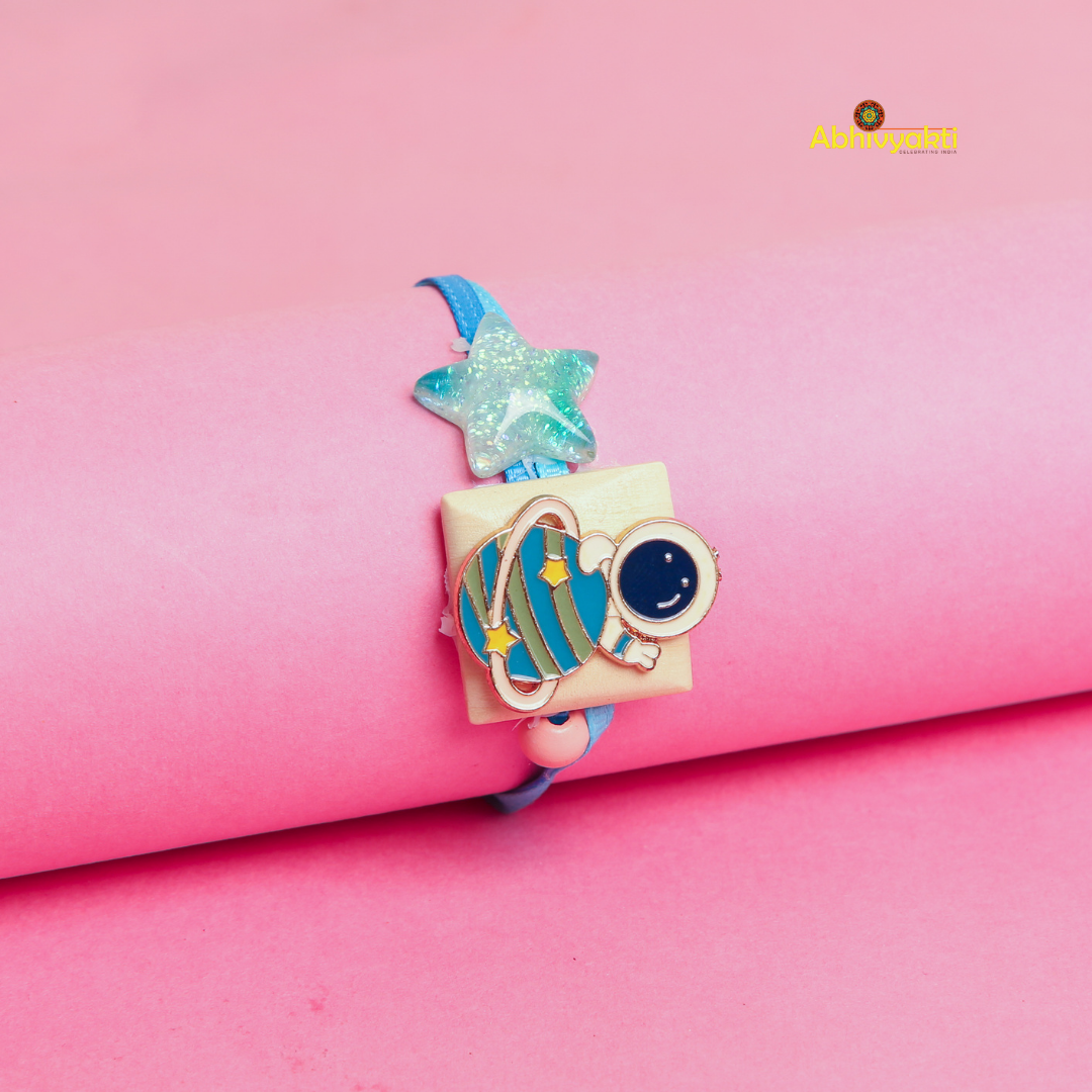 Cartoon astronaut and planet Rakhi bracelet for kids, featuring a blue strap, a glittery star, and a cute astronaut with a planet charm, displayed on a pink background.