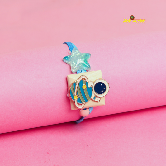 Cartoon astronaut and planet Rakhi bracelet for kids, featuring a blue strap, a glittery star, and a cute astronaut with a planet charm, displayed on a pink background.