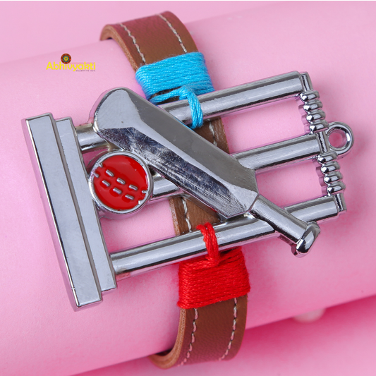 A Kids bracelet with a cricket-themed charm is displayed against a pink background. The charm features a bat, ball, and stumps in a metallic finish, while the bracelet itself is brown leather with blue and red thread accents—an ideal rakhi for kids who love the game.