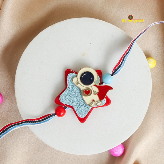 A colorful rakhi with a star-shaped base and an astronaut design, placed on white and golden surface. The rakhi has blue, red, and white threads and beads. 