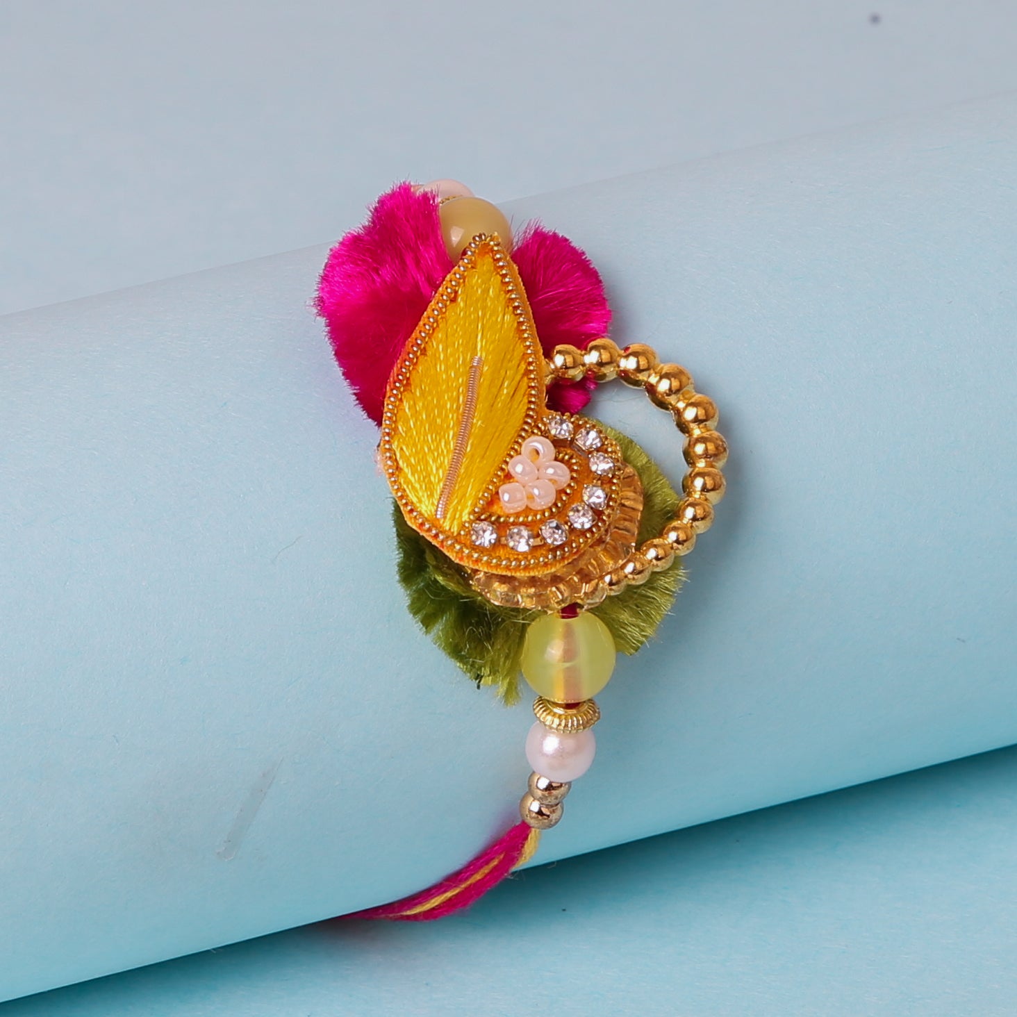  A decorative rakhi with a yellow and gold centerpiece, pink and green accents, pearls, golden beads, and stones and scattered pearls