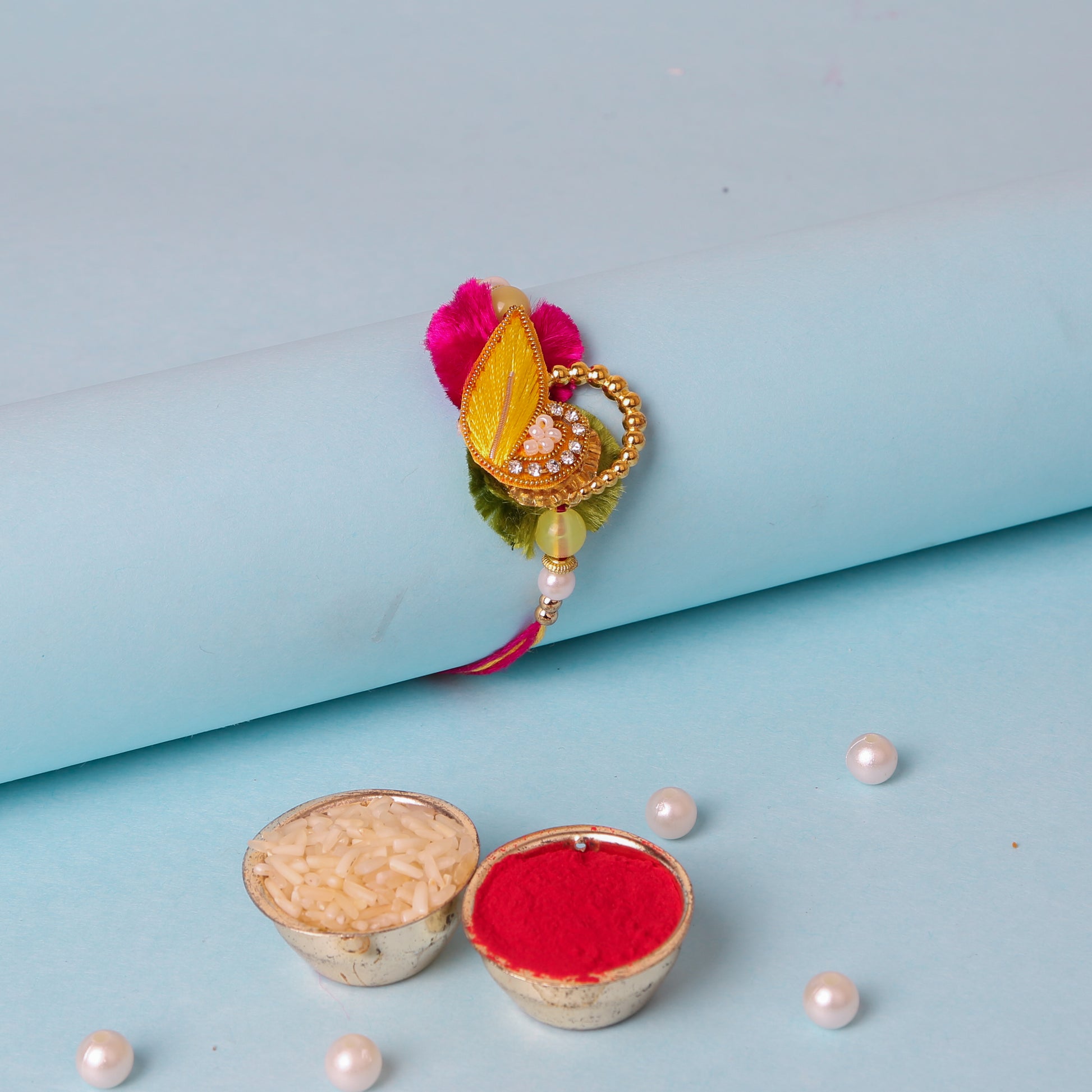  A decorative rakhi with a yellow and gold centerpiece, pink and green accents, pearls, golden beads, and stones, placed on a blue background with bowls of rice and kumkum, and scattered pearls