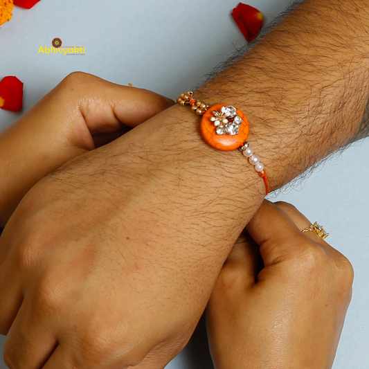 A decorative rakhi with an orange circular center adorned with crystals, small gold beads, and white pearls, attached to an orange thread, displayed on a light blue background.