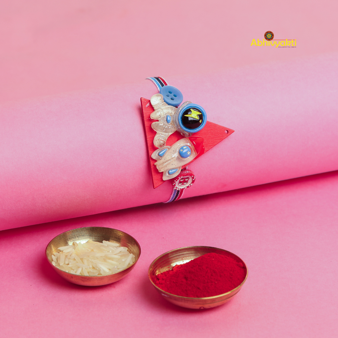 Baby Astronaut Rakhi bracelet for kids, with a cute cartoon astronaut and spaceship charm on a multicolor thread,with two bowls one rice and one with roli, placed on a pink background