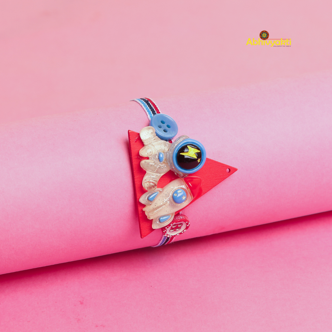 Baby Astronaut Rakhi bracelet for kids, with a cute cartoon astronaut and spaceship charm on a multicolor thread, placed on a pink background.