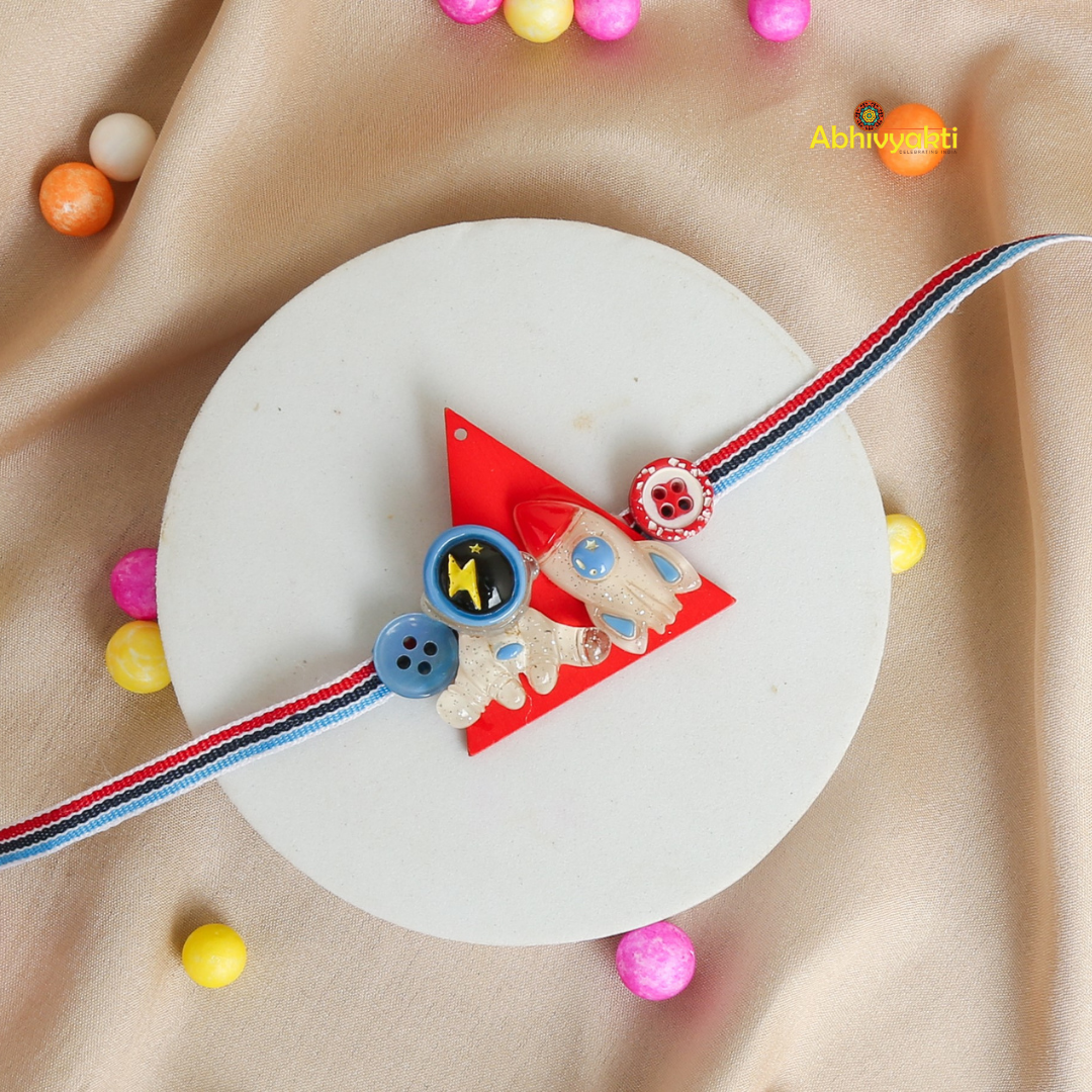 Baby Astronaut Rakhi bracelet for kids, with a cute cartoon astronaut and spaceship charm on a pastel green thread, placed on a beige background with colorful balls around it.