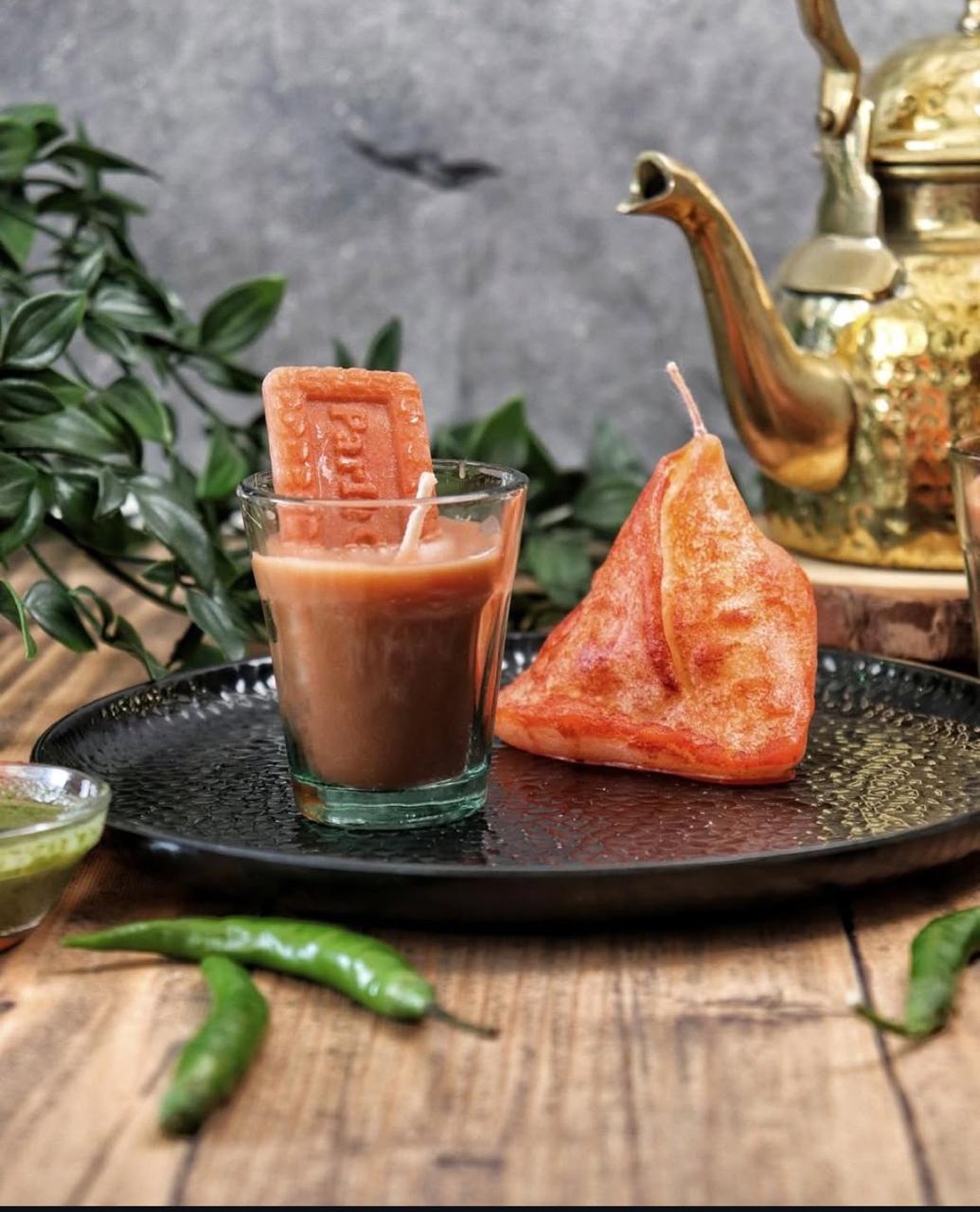 Candle shaped like a Samosa with a glass of Cutting chaion a dark tray, with a teapot and green leaves in the background.