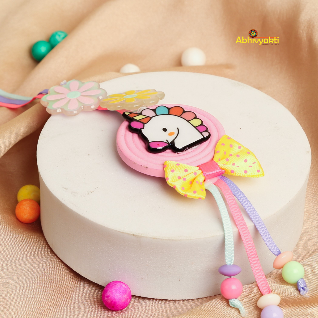 A colorful rakhi lumba with a unicorn design, two flower decorations, a yellow polka-dot bow, and pastel beads, displayed on  golden and white background.
