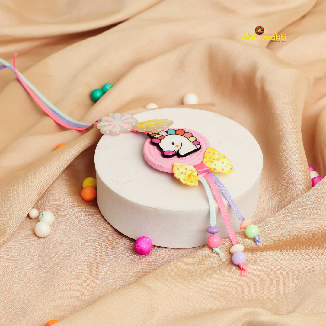 A colorful rakhi lumba with a unicorn design, two flower decorations, a yellow polka-dot bow, and pastel beads, displayed on golden  background.