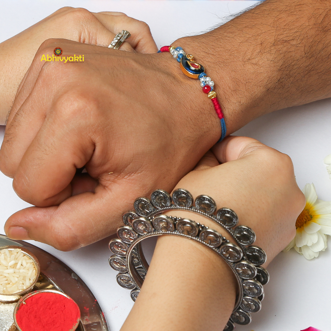 A decorative bracelet with red and blue threads wrapped around a cylindrical light blue object. The centerpiece is a gold emblem featuring a blue teardrop shape with a red inset, reminiscent of the intricate designs found in kundan paisley rakhi, surrounded by small beads in various colors.