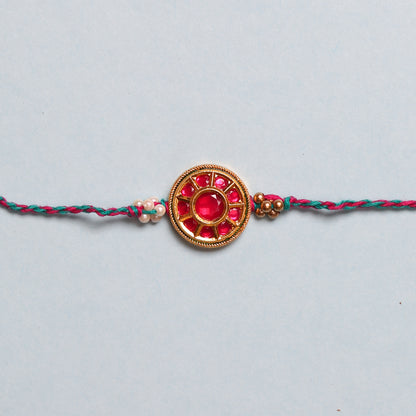 Elegant rakhi with a circular gold centerpiece, bright red stones forming a sunburst pattern, and teal-pink braided thread with pearl and gold beads, on a light blue background. 