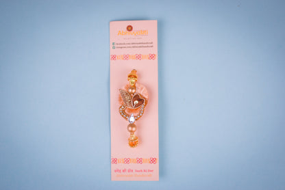 Fancy rakhi featuring a gold and brown leaf motif with sparkling stones, beige pom-pom, hanging bead, placed on a blue roll with rice, roli, and a pearl bracelet nearby