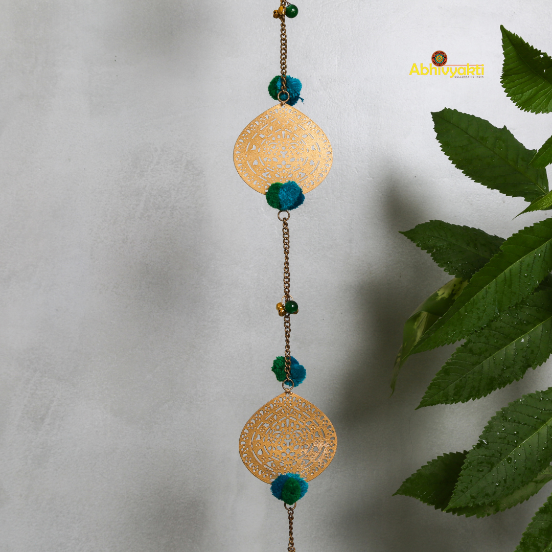 Filigree metal door hanging in a circular shape featuring detailed ornamental design, adorned with blue pom-poms and tiny golden bells, suspended from a chain indoors 
