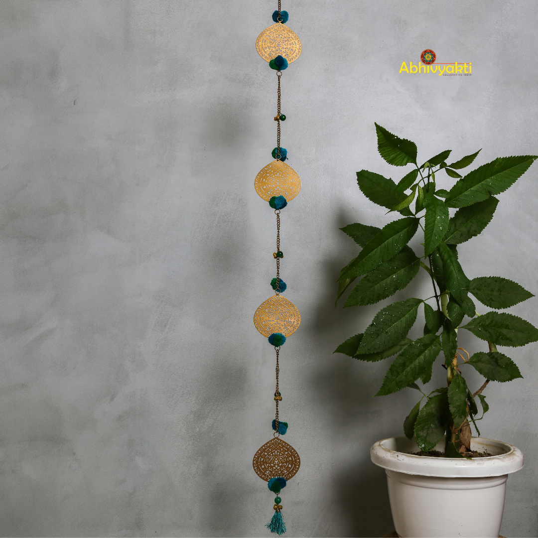 Filigree metal door hanging in a circular shape featuring detailed ornamental design, adorned with blue pom-poms and tiny golden bells, suspended from a chain indoors 