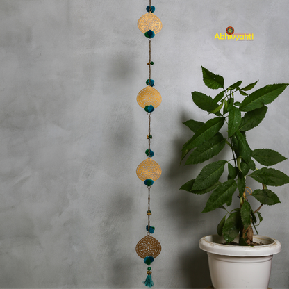 Filigree metal door hanging in a circular shape featuring detailed ornamental design, adorned with blue pom-poms and tiny golden bells, suspended from a chain indoors 